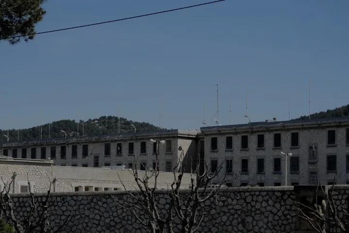 Edificio carcerario in pietra osservato oltre una recinzione scura sotto un cielo livido